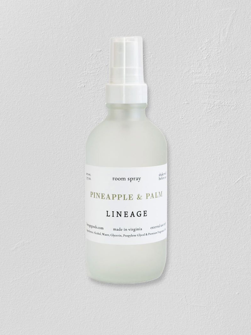A frosted glass room spray bottle with a white spray top and a white label reading 'PINEAPPLE & PALM LINEAGE' on a light gray textured background.